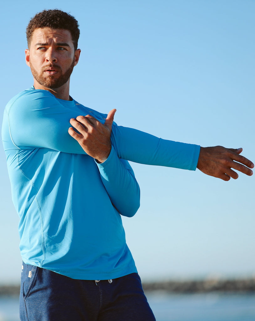 Man stretching at the beach wearing long sleeve jet tee shirt in ocean blue. (Style 12002) - BloqUV