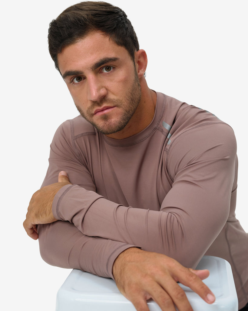Man wearing a brown long-sleeve shirt sitting on a white stool against a plain background