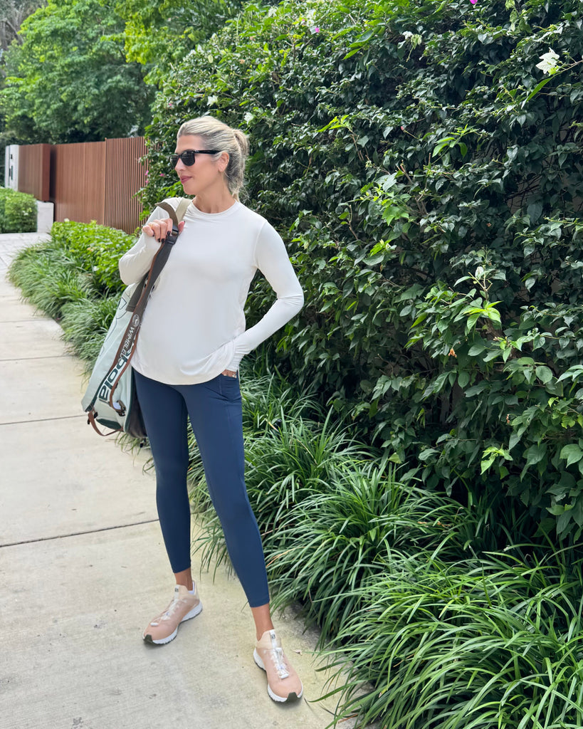 Woman in white top and blue leggings standing on a sidewalk with greenery in the background
