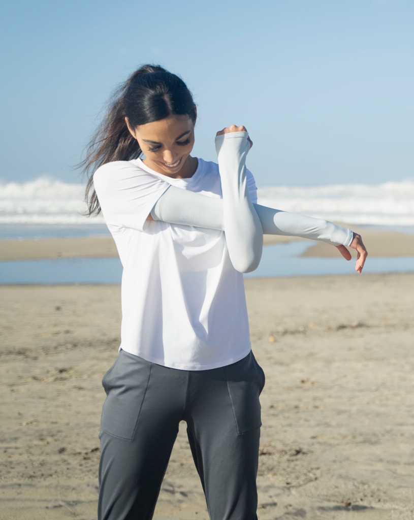 Women stretching at the beach wearing white short sleeve crew top with soft grey sun sleeves and smoke joggers. (Style 1101) - BloqUV