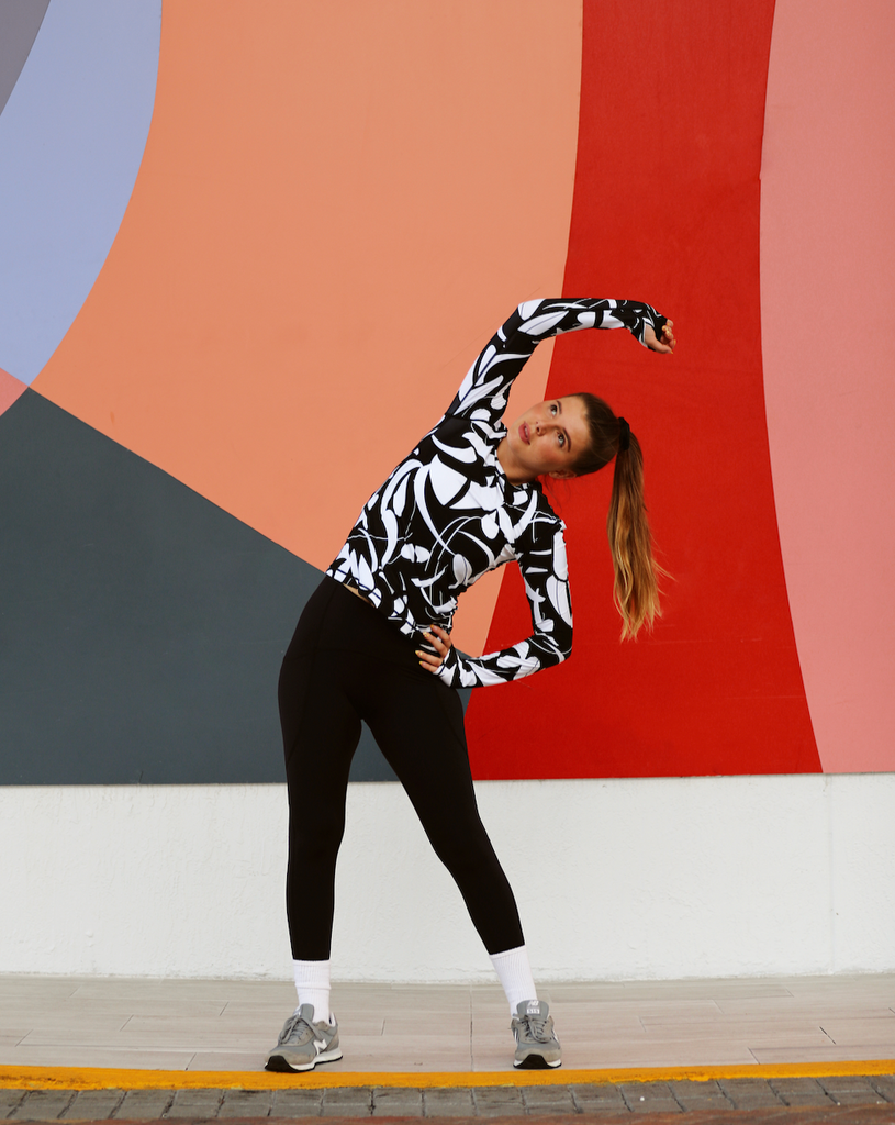 Women stretching outdoors in front of colorful wall wearing printed black and white long sleeve 24/7 shirt with black tights. (Style 2001J) - BloqUV
