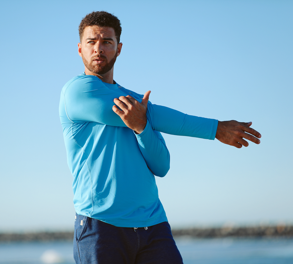 Man wearing long sleeve jet tee in ocean blue while stretching at the beach.