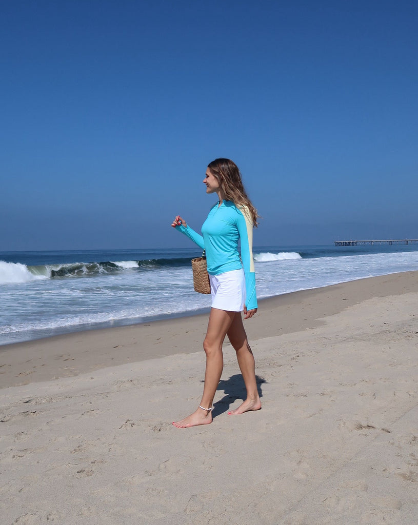 Woman in turquoise top and white shorts standing on a sandy beach with ocean waves and clear blue sky.