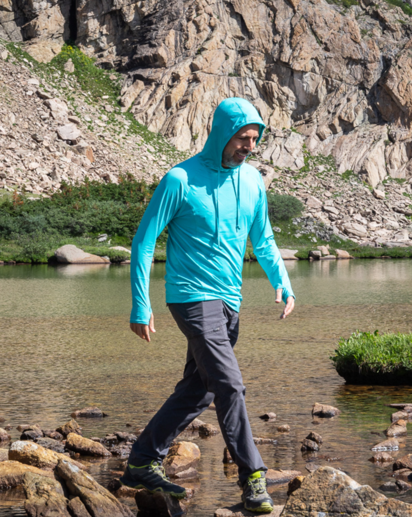 Man walking over rocks in river wearing caribbean blue color unisex long sleeve hoodie shirt. Hoodie is over his head.(Style 12007) - BloqUV