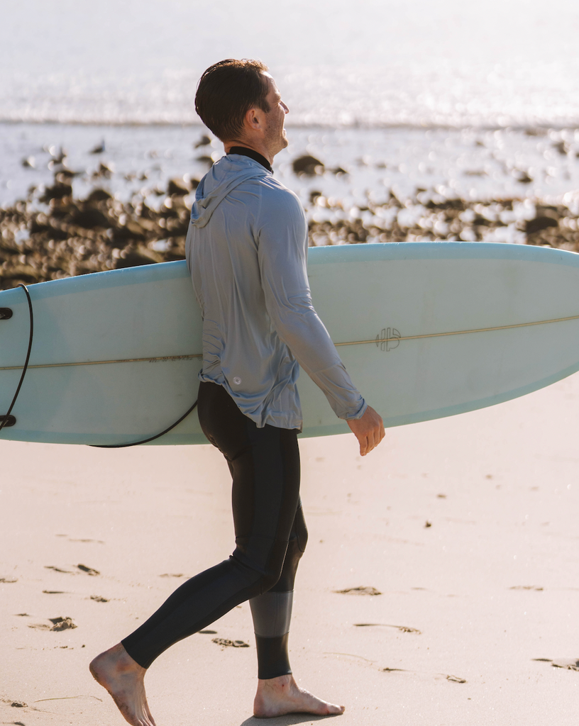 Man holding surf board on the beach wearing soft grey unisex long sleeve hoodie shirt.  (Style 12007) - BloqUV